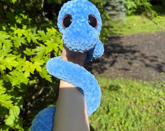 May include: A bright blue plush snake toy made of soft, textured yarn. The snake features large, round, black safety eyes with subtle pink glitter accents around the edges. The long, flexible body is coiled, showcasing a thick, fuzzy construction.