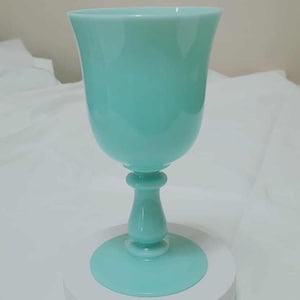 Puede incluir: Una sola copa de color verde azulado claro. El vaso tiene un cuenco redondeado, un tallo delgado y una base circular. La copa está hecha de un material sólido y opaco y se exhibe sobre una superficie blanca.