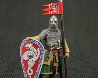 Puede incluir: Figura en miniatura de un caballero medieval, vestido con cota de malla y sosteniendo un escudo con un emblema de dragón. El caballero también sostiene una lanza y una bandera roja con un emblema blanco. La figura está sobre una base pequeña.