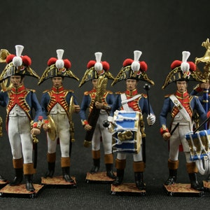 May include: A set of eight miniature toy soldiers in military uniforms, playing instruments and holding drums. The soldiers are dressed in blue and white uniforms with red accents and white feathered hats. The figures are posed in a marching band formation.
