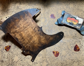 Peut inclure: Un pickguard de guitare avec un dégradé de brun foncé et de noir et une finition texturée, comportant plusieurs petits trous de montage. Trois médiators à motif écaille sont dispersés à proximité, avec une petite carte de visite.
