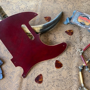 May include: A burgundy guitar pickguard with a cutout for the pickups and controls, along with several tortoiseshell guitar picks. A decorative necklace and a business card are also present.