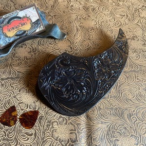 Puede incluir: Una púa de guitarra de cuero negro con un diseño floral en relieve. La púa tiene forma de lágrima y un acabado liso y pulido.