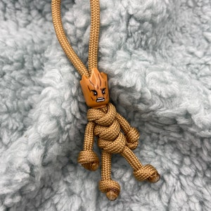 May include: A brown paracord keychain with a knotted figure of a character with a brown head and a grumpy expression. The keychain is on a light blue fluffy surface.