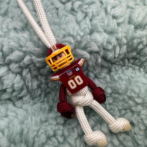 Könnte beinhalten: Weißer Paracord-Schlüsselanhänger mit einem roten, weißen und goldenen Fußballspieler-Anhänger. Der Spieler trägt einen Helm und ein Trikot mit der Nummer "00" und "49" darauf.