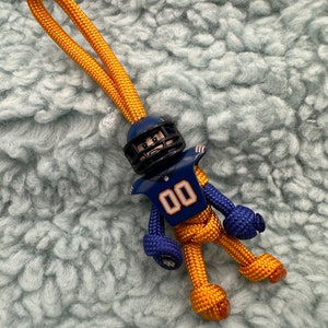 Könnte beinhalten: Ein gold-blauer Paracord-Schlüsselanhänger mit einer Fußballspieler-Figur. Die Figur trägt einen Helm und ein Trikot mit der Nummer "00" darauf.