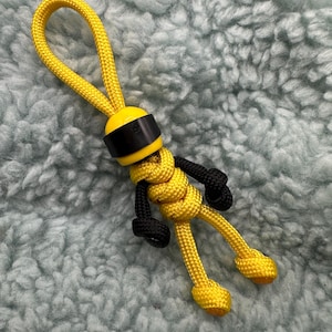 Könnte beinhalten: Ein handgefertigter gelb-schwarzer Paracord-Schlüsselanhänger in Form einer kleinen Figur. Die Figur hat einen gelben Kopf mit einem schwarzen Band, schwarze Arme und Beine und einen gelben Körper. Die Schlüsselanhängerschlaufe ist ebenfalls gelb.