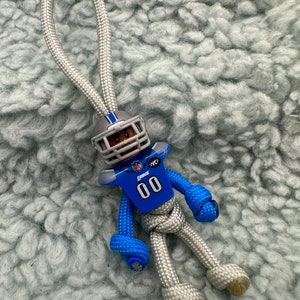 Könnte beinhalten: Ein silberner und blauer Paracord-Schlüsselanhänger mit einem Miniatur-Footballspieler, der ein Trikot der Detroit Lions mit der Nummer 00 trägt. Der Schlüsselanhänger hat oben eine Schlaufe zum Anbringen an einer Tasche oder Schlüssel.
