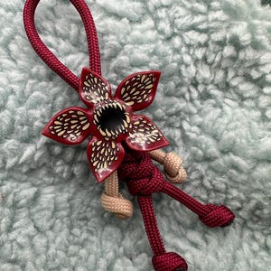 Könnte beinhalten: Ein roter und beiger Paracord-Schlüsselanhänger mit einem roten und gelben Blumenanhänger, der einen schwarzen Mund mit Zähnen zeigt. Der Blumenanhänger ist eine Darstellung des Demogorgons aus der Serie Stranger Things.
