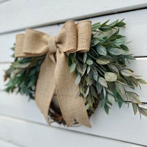 May include: A rustic wreath made of green leaves with a large burlap bow. The wreath is hanging on a white wooden wall.
