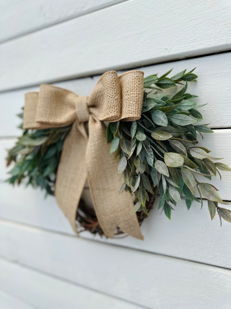 May include: A rustic wreath made of green leaves with a large burlap bow. The wreath is hanging on a white wooden wall.