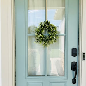 May include: A light blue front door with a green wreath hanging on it. The door has a glass pane with a white curtain behind it. There is a black door handle and a black keypad on the right side of the door.