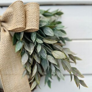 May include: A burlap ribbon bow tied around a green leafy wreath. The wreath is made of artificial greenery and is perfect for a farmhouse or rustic decor style.