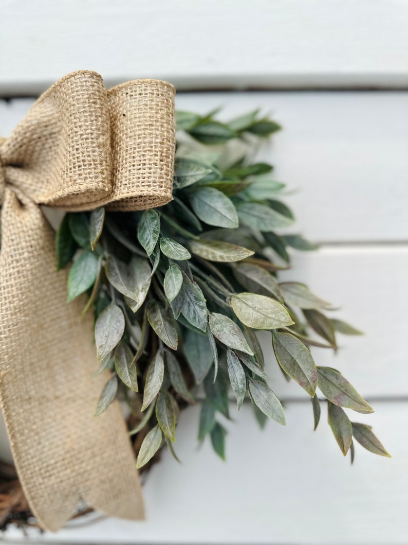 May include: A burlap ribbon bow tied around a green leafy wreath. The wreath is made of artificial greenery and is perfect for a farmhouse or rustic decor style.