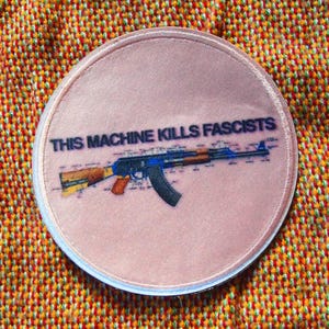 May include: A circular patch featuring the text "THIS MACHINE KILLS FASCISTS" and a blue and brown rifle illustration. The patch is displayed on a textured orange and yellow fabric.