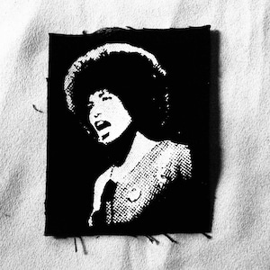 Può includere: Immagine in bianco e nero di una donna con un'acconciatura afro. Indossa una camicia scura e sta parlando. L'immagine è su un cerotto di stoffa nero.