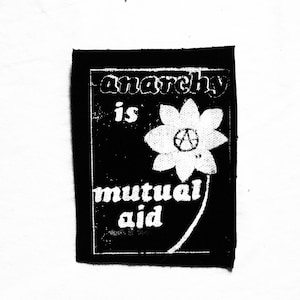 May include: Black and white patch with the text "anarchy is mutual aid". A white flower with a peace symbol in the center is in the middle of the patch. The patch is rectangular with rounded corners.