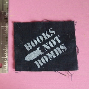 Puede incluir: Un parche de tela negro con texto blanco que dice "BOOKS NOT BOMBS" y una silueta blanca de un avión.