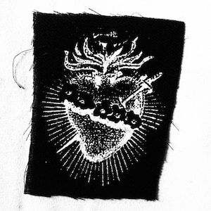 May include: Black square patch featuring a white Sacred Heart design. The heart is surrounded by a radiant burst of light, with flames at the top and a sword piercing the heart. The word "Sacred" is visible.