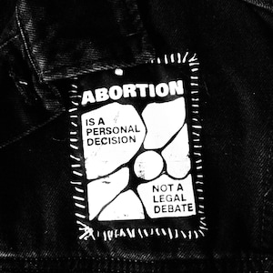 May include: Black and white patch sewn onto a black denim jacket. The patch reads "ABORTION IS A PERSONAL DECISION NOT A LEGAL DEBATE".