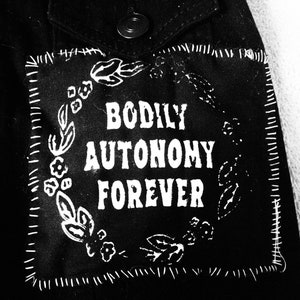 Puede incluir: Chaqueta vaquera negra con un parche blanco que dice "Bodily Autonomy Forever" rodeado de un diseño floral.