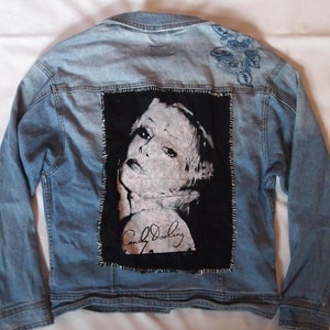 May include: A light blue denim jacket with a black patch featuring a photo of a woman with the signature "Candy Darling" on it. The jacket has a floral embroidery on the upper back.