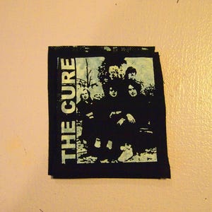 Op de afbeelding: Zwart-witte patch met de tekst "THE CURE" in witte letters. De patch toont een silhouet van een band met vijf leden.