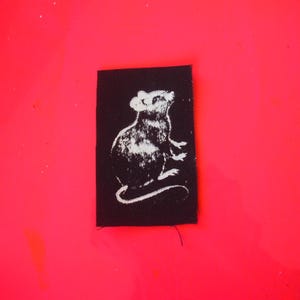 May include: A black rectangular patch featuring a white illustration of a rat. The rat is depicted in a stylized, upward-facing pose. The patch is set against a bright red background.