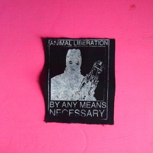 以下が含まれることがあります： 「ANIMAL LIBERATION」と「BY ANY MEANS NECESSARY」というテキストが書かれた白黒のパッチ。パッチには、マスクを着用し、道具を持っている人が描かれている。