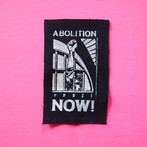 Puede incluir: Parche blanco y negro con las palabras "Abolition Now!" y un puño en alto rompiendo una celda de prisión.