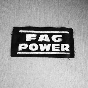 Peut inclure: Écusson en tissu noir avec du texte blanc qui dit "FAG POWER".