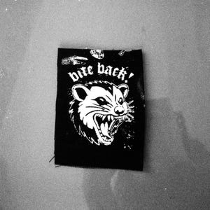 Può includere: Toppa nera e bianca con un opossum ringhiante e la scritta "bite back!". La toppa rettangolare è progettata per essere cucita o stirata su vestiti o borse.