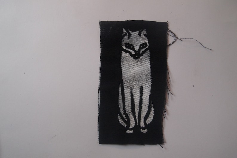 May include: A black fabric patch with a white silhouette of a cat. The cat is facing forward with its tail behind it.