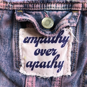 May include: A close-up of a denim jacket with a pink and blue faded wash. A white patch is sewn onto the jacket with the words "empathy over apathy" in dark blue lettering.