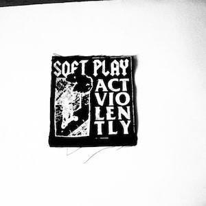 Könnte beinhalten: Schwarz-weiß-Patch mit dem Text "Soft Play Act Violently" in fetter gotischer Schrift. Der Patch zeigt eine Grafik einer Person, die von einem Gebäude springt.