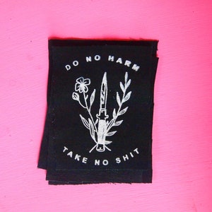 Może przedstawiać: Czarna łatka z tkaniny z białym haftem. Łatka głosi "Do No Harm Take No Shit" i przedstawia nóż z kwiatami.