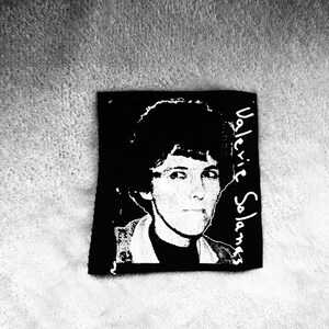 May include: Black and white image of a person with short dark hair. The person is wearing a dark shirt and has the name "Valerie Solanas" written in white text on the right side of the image.