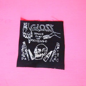 May include: Black iron-on patch with white text and graphics. The text reads "GLOSS TRANS DAY OF REVENGE". The graphics include two hands, a skull, and several knives.
