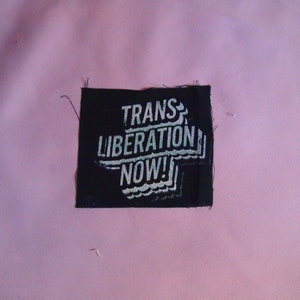 Puede incluir: Parche de tela negro con texto blanco que dice "TRANS LIBERATION NOW!" en una fuente estilizada.