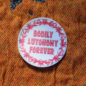 Puede incluir: Un parche redondo blanco con las palabras "BODILY AUTONOMY FOREVER" en rojo, rodeado por un diseño floral y de hojas rojas. El parche se encuentra sobre un fondo naranja y marrón texturizado.