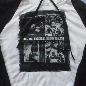 Puede incluir: Sudadera con capucha raglán blanca y negra con un parche negro cosido en la parte delantera. El parche presenta cuatro imágenes en blanco y negro y el texto "ALL YOU FASCISTS BOUND TO LOSE".