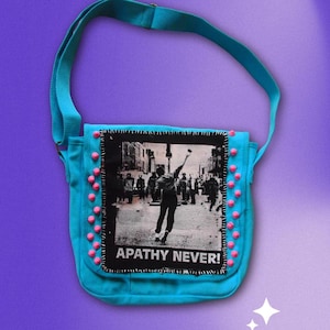 May include: A turquoise messenger bag with a black and white graphic print of a person throwing an object, with the text "APATHY NEVER!". The bag has a turquoise strap and is decorated with pink pom-poms.