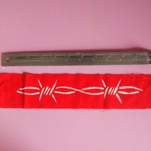 Puede incluir: Un parche de tela rojo con un diseño de alambre de púas blanco. El parche mide aproximadamente 10 cm de largo y 4 cm de ancho.