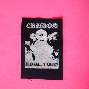 May include: Black and white patch with the text "CRUDOS" and "ILEGAL, Y QUE?" in a gothic font. The patch features a silhouette of a person with a hood.