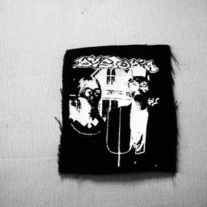 Op de afbeelding: Zwart-witte geborduurde patch met de tekst "Svastika" en een afbeelding van twee figuren met gasmaskers voor een huis.