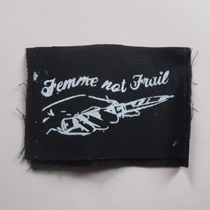 Może przedstawiać: Czarna łatka z tkaniny z białym tekstem i grafiką dłoni trzymającej nóż. Tekst brzmi "Femme not Frail".