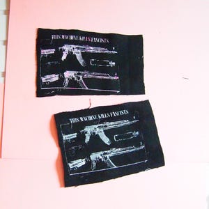 Può includere: Due toppe di tessuto nero con una grafica bianca e rosa di un fucile e la scritta "THIS MACHINE KILLS FASCISTS". Le toppe sono rettangolari e hanno un aspetto leggermente usurato.