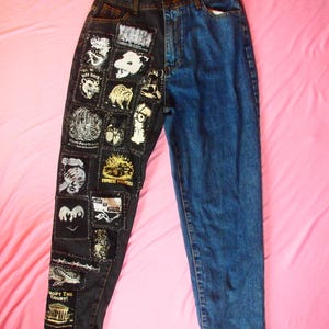 Puede incluir: Vaqueros de mezclilla azules con un diseño único. Una pierna es azul liso, mientras que la otra es negra y está cubierta de parches con varias ilustraciones y texto. Los parches incluyen imágenes de animales, caras y frases como "EXPRESS YOURSELF".
