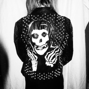May include: Black leather jacket featuring a white skull graphic on the back. The jacket is studded with silver metal studs around the collar and shoulders. The skull graphic is a stylized design with a grinning mouth and skeletal hands.