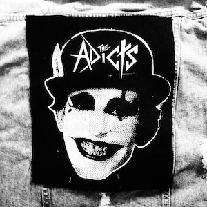 May include: Black and white patch showing a smiling face in a bowler hat, with the words "The Adicts" above. The patch is attached to a denim jacket. The image is in monochrome.